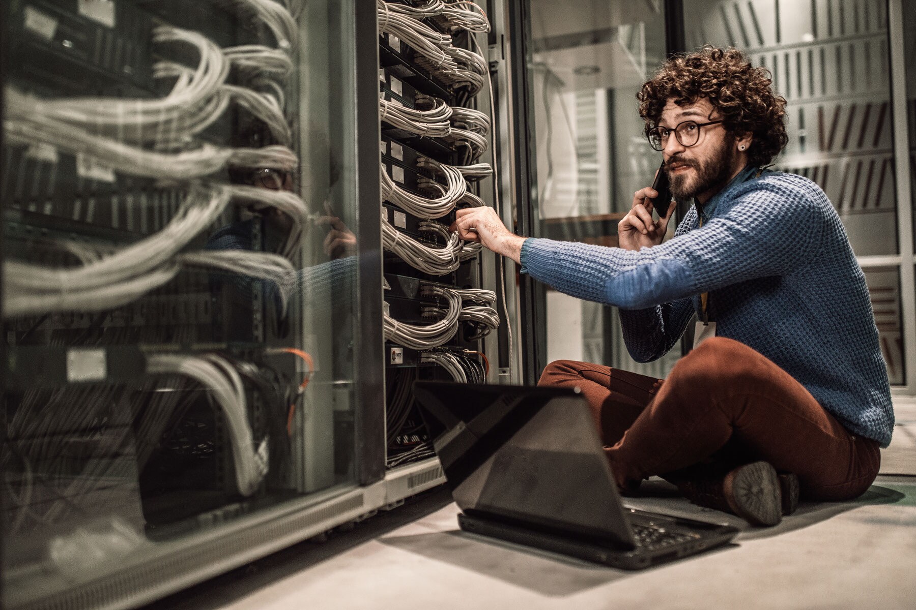 Person sitting on the floor next to a server inside a data centre while talking to someone on the phone and has his laptop on his lap.